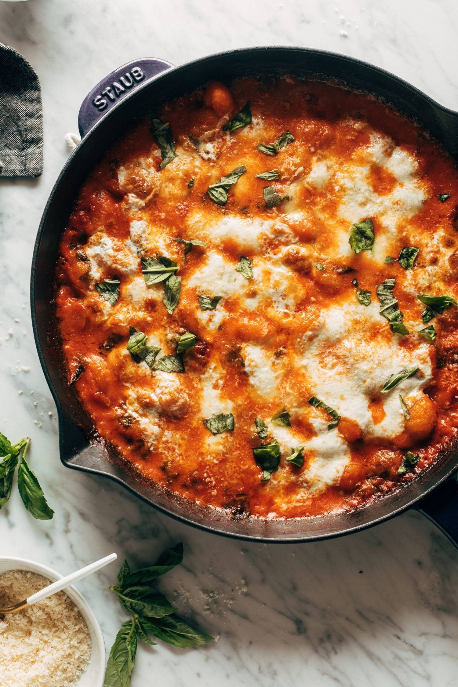 Three cheese baked gnocchi sprinkled with fresh basil on top. The gnocchi is in a cast iron pan and there's fresh basil and grated parmesan cheese in a bowl on the side.